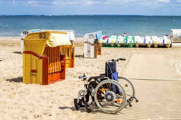 Rollstuhl steht auf Sandmatte am Strand bei Strandkörben mit Meer im Hintergrund
